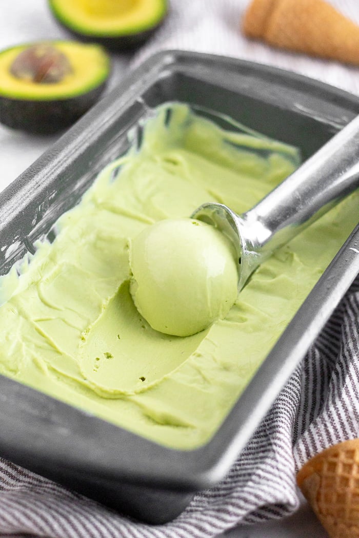 Close up of an ice cream scoop scooping avocado frozen yogurt out of a loaf pan with avocados and ice cream cones around it.