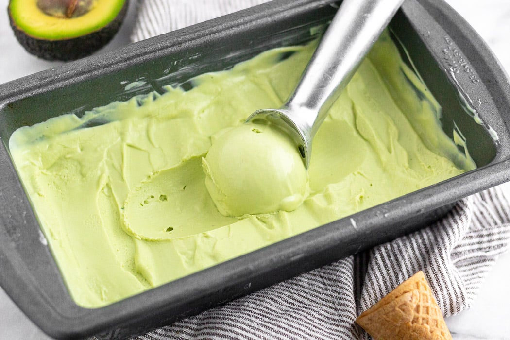 Loaf pan filled with avocado frozen yogurt with a ice cream scoop taking some out of it. It is surrounded by an avocado and an ice cream cone.