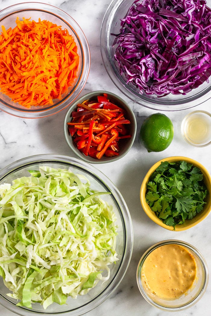 Different bowls filled with carrots, shredded purple cabbage, red peppers, cilantro, green cabbage, chipotle mayo, apple cider vinegar, and a lime.