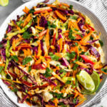Large bowl of chipotle Mexican coleslaw with lime wedges on the side of the bowl. It is stilling on a stripped towel with lime wedges and cilantro around it.