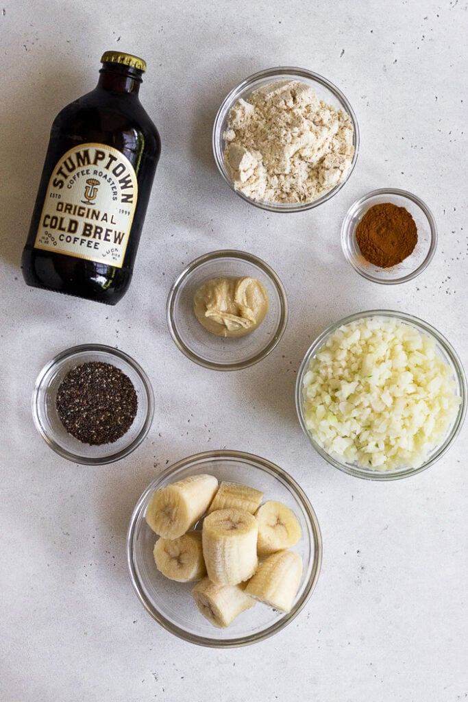 Overhead shot of a bottle of cold crew coffee, bowl of protein powder, bowl of cinnamon, bowl of cauliflower rice, bowl of frozen bananas, bowl of chia seeds, and a bowl of cashew butter.