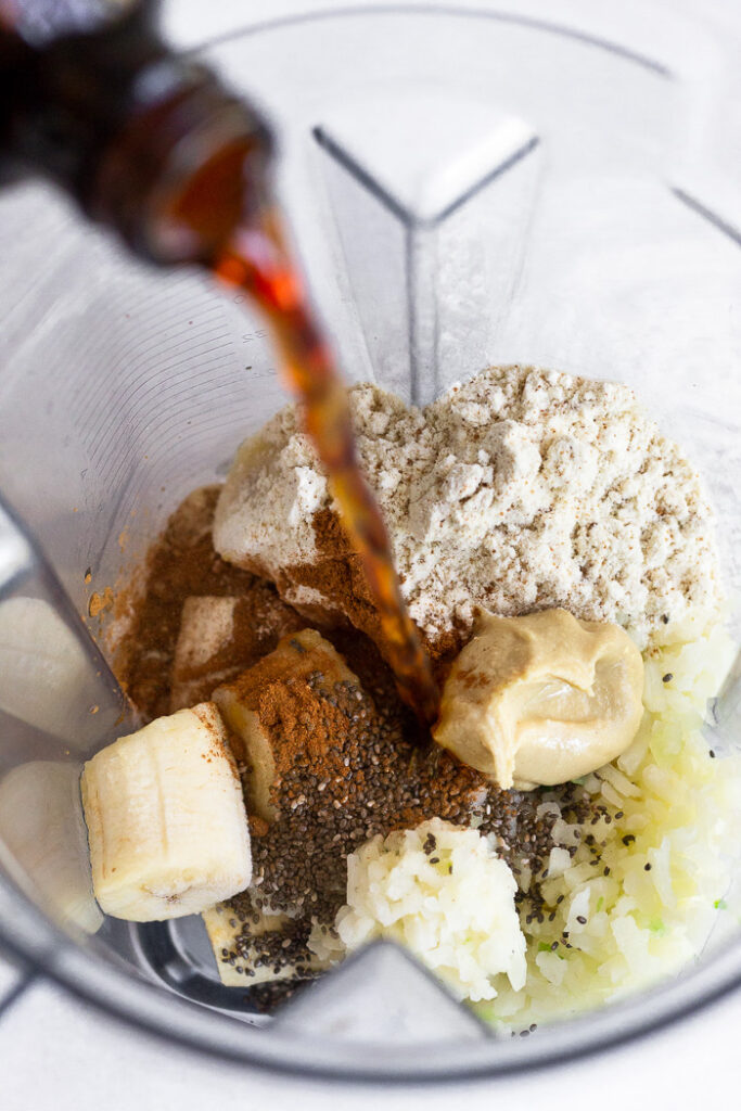 Blender filled with frozen bananas, cauliflower rice, protein powder, cashew butter, chia seeds, and cinnamon with cold brew being poured into it.