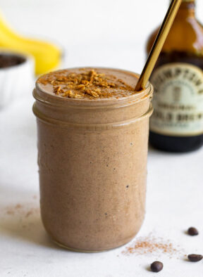 Coffee protein smoothie in a jar with some toppings on it and a straw in it. Around it is a jar of cold brew, coffee beans, and bananas.