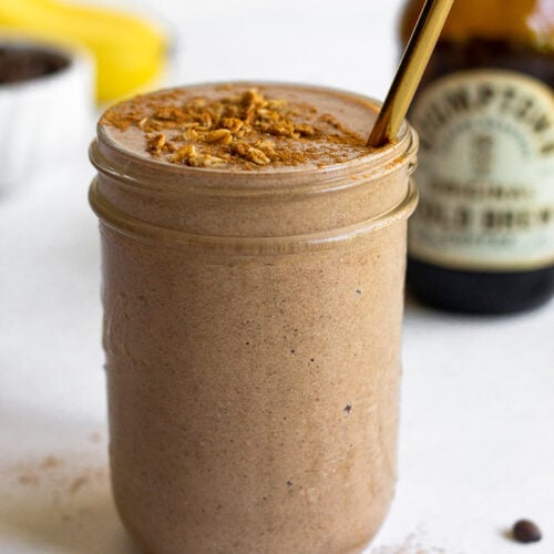Coffee protein smoothie in a jar with some toppings on it and a straw in it. Around it is a jar of cold brew, coffee beans, and bananas.