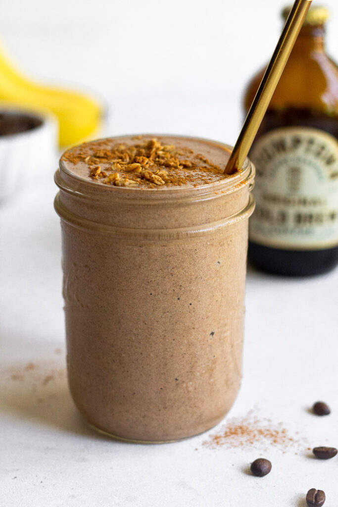 Coffee protein smoothie in a jar with some toppings on it and a straw in it. Around it is a jar of cold brew, coffee beans, and bananas.