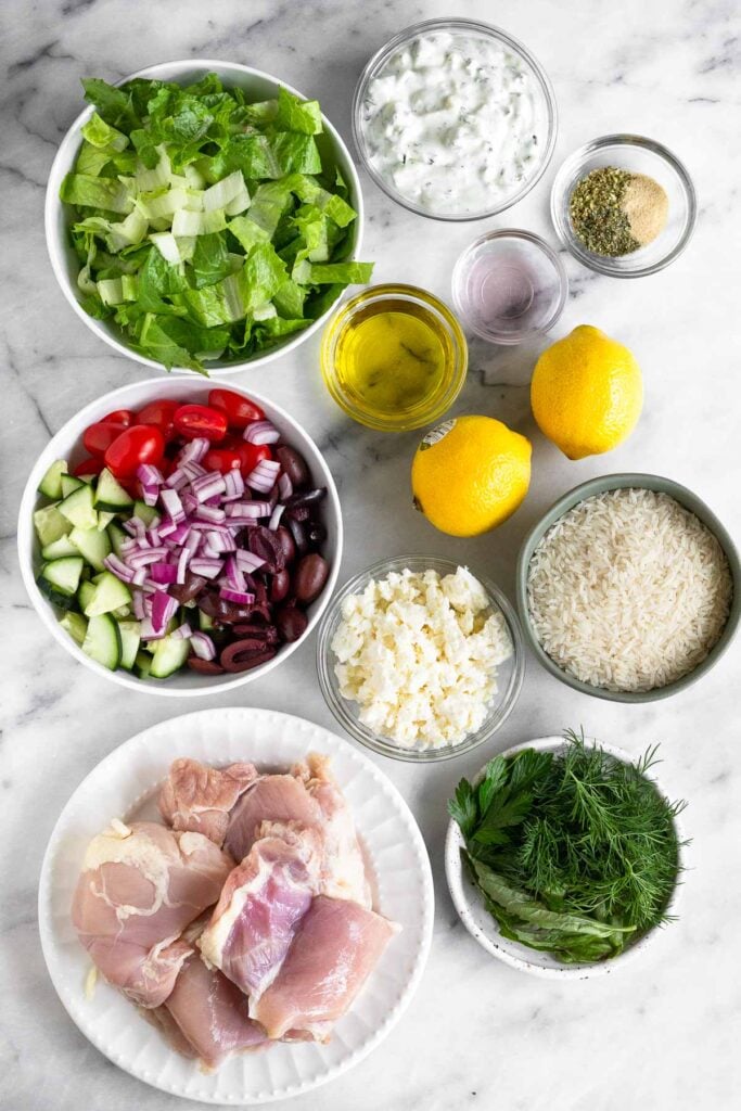 Marble counter with a bowl of tzatziki sauce, a bowl of spices, a bowl of red wine vinegar, 2 lemons, a bowl of uncooked white rice, a small plate of fresh herbs, a bowl of feta, a plate of raw chicken thighs, a bowl of chopped tomatoes, cucumber, and olives, a bowl of olive oil, and a bowl of chopped romaine.