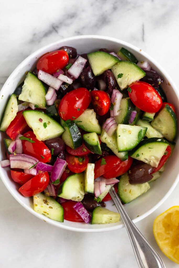 Greek salad in a bowl with chopped tomatoes, diced cucumbers, olives, red onion, and tossed with lemon, oil, and spices. A spoon is in the bowl and next to the bowl is a half a juiced lemon.