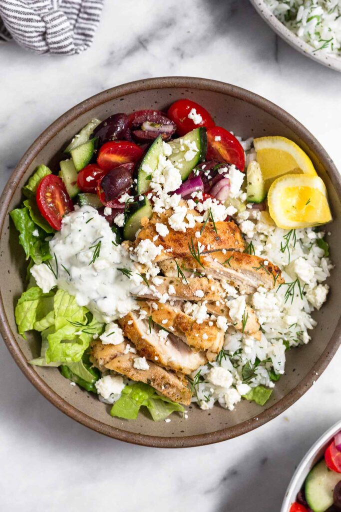 Greek bowl with chopped romaine, diced tomatoes, cucumbers, and olives, herby white rice, sliced chicken thighs, tzatziki, and feta and garnished with dill and 2 lemon wedges. Next to the bowl is a bowl of tomato cucumber salad, a bowl of herby rice, and a kitchen towel.