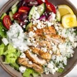 Greek chicken bowl with chopped romaine, tomato cucumber salad, herby rice, sliced chicken thighs, tzatziki, and feta and garnished with dill and 2 lemon wedges.