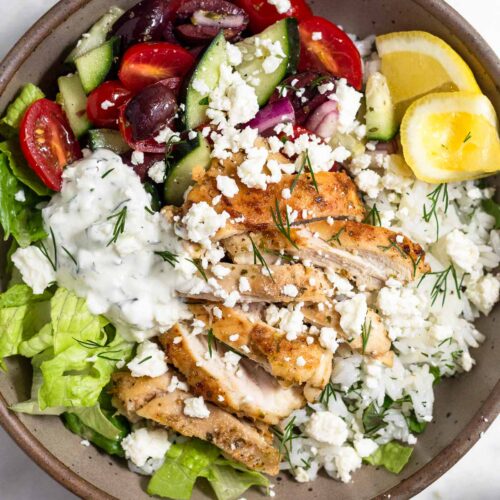 Greek chicken bowl with chopped romaine, tomato cucumber salad, herby rice, sliced chicken thighs, tzatziki, and feta and garnished with dill and 2 lemon wedges.