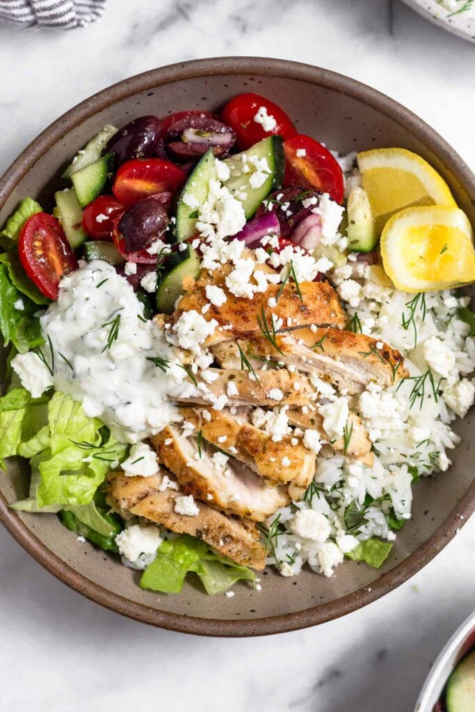 Greek chicken bowl with chopped romaine, tomato cucumber salad, herby rice, sliced chicken thighs, tzatziki, and feta and garnished with dill and 2 lemon wedges.