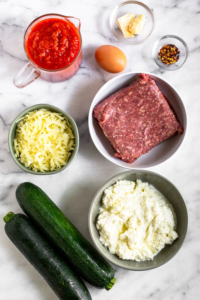 White marble counter top with pasta sauce, an egg, red pepper flakes, ground beef, ricotta cheese, parmesan cheese, and 2 zucchinis.