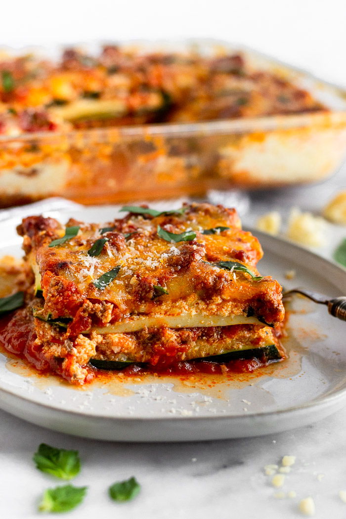 Slice of low carb zucchini lasagna on a white plate with a fork next to it. It is garnished with fresh basil and small basil leaves around the plate. Behind it is pan of more zucchini lasagna.