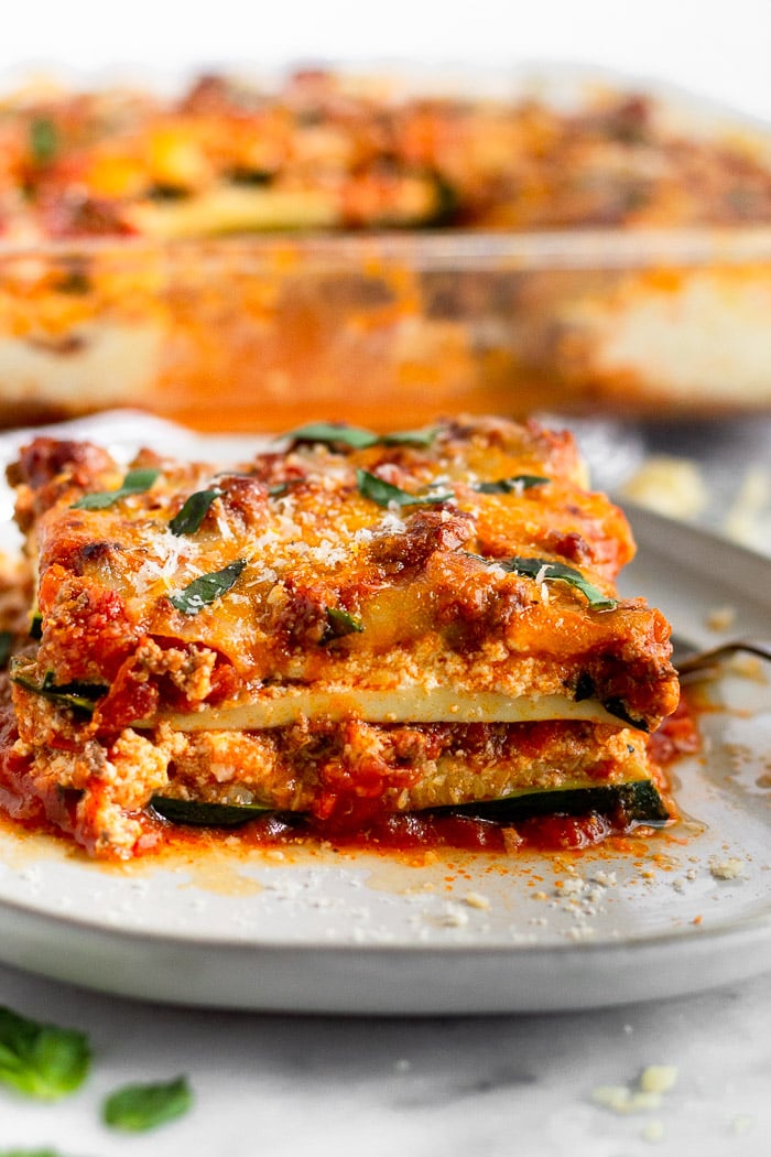 White plate with a slice of low carb zucchini lasagna topped with parmesan cheese and fresh basil. Behind it is a baking dish of more zucchini lasagna,
