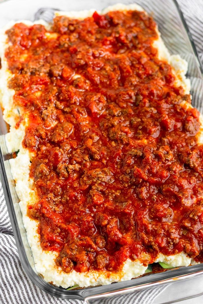 The making of zucchini lasagna. It is in a large glass baking dish with a layer of cheese and then meat sauce.