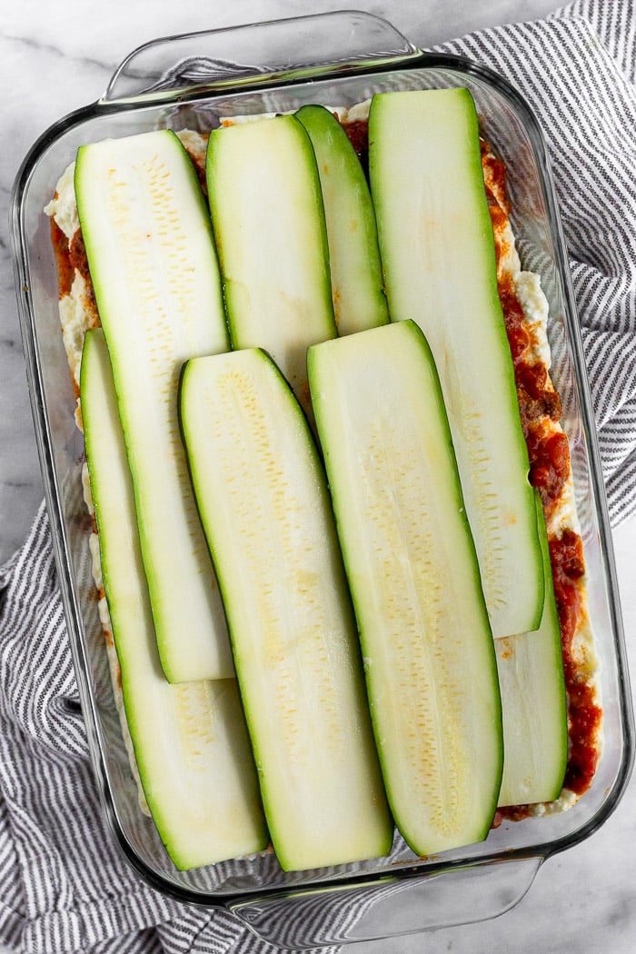 The making of zucchini lasagna. It is in a large glass baking dish with a layer of cheese, then meat sauce, and finally zucchini slices.
