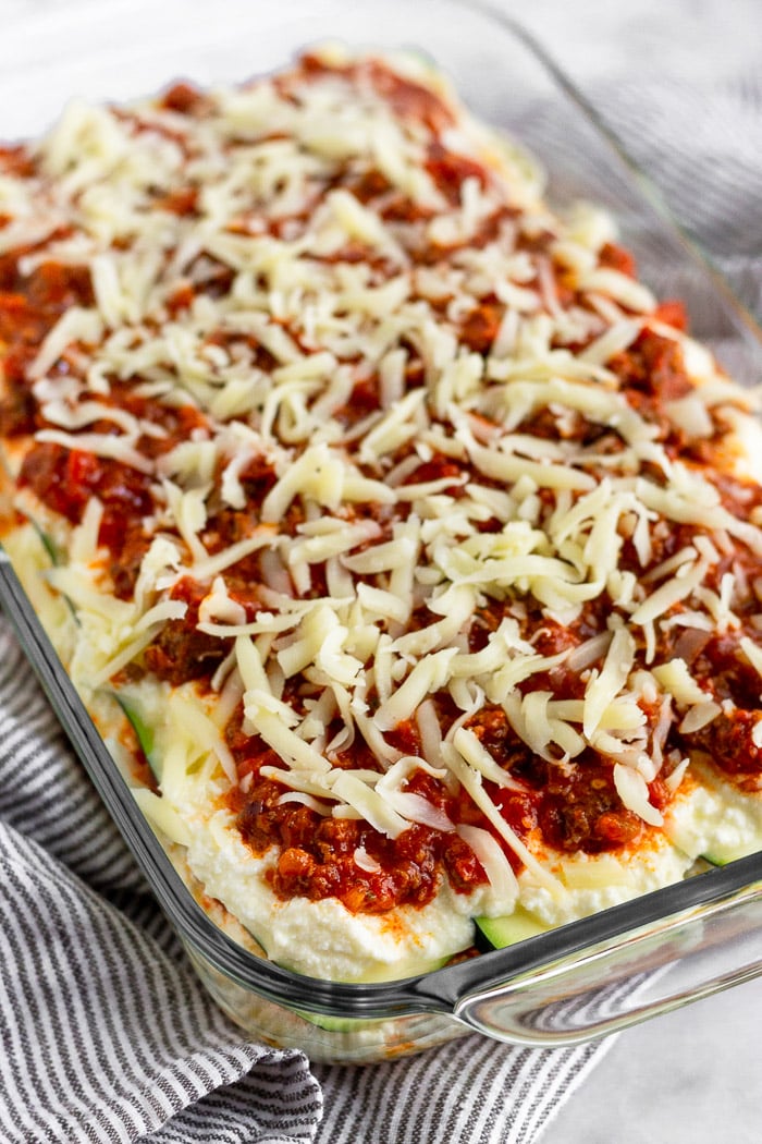 Low carb zucchini lasagna before it goes into the oven. It has layers of beef, meat sauce, zucchini, and cheese and the top is sprinkled with more cheese.