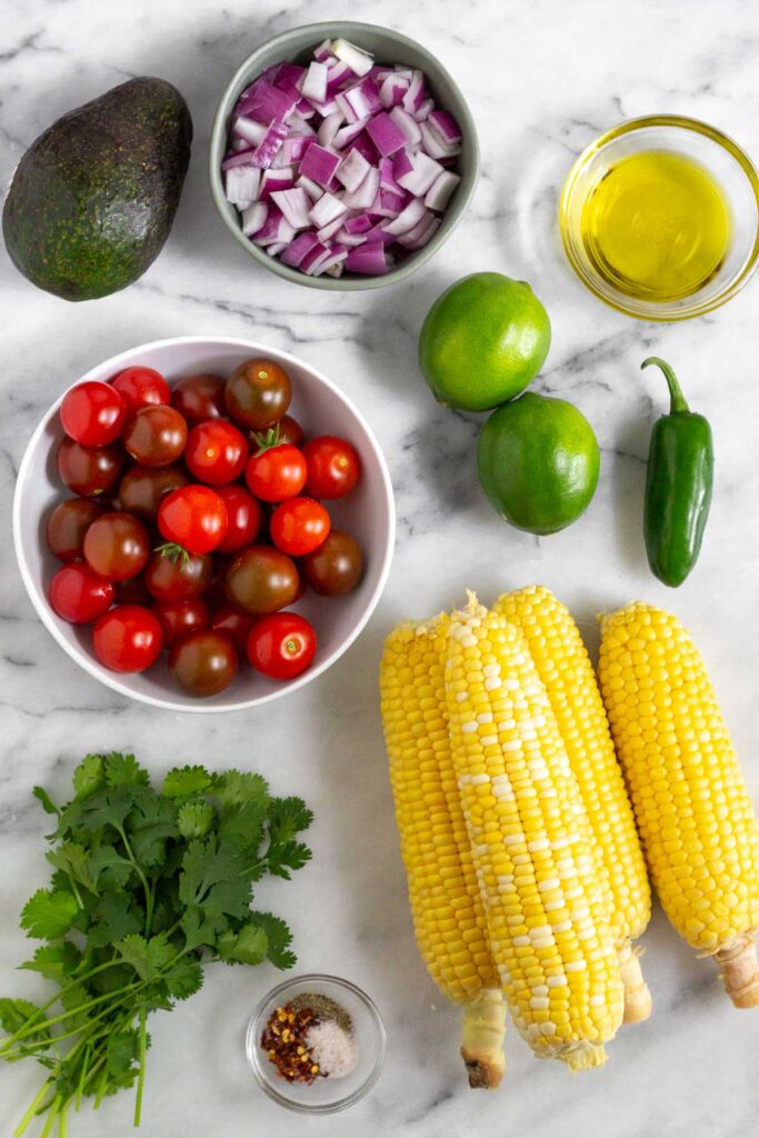 White marble counter top with a bowl of olive oil, 2 limes, a jalapeño, 4 ears of corn, a bowl of spices, some cilantro, a bowl of cherry tomatoes, a whole avocado, and a bowl of diced red onion.