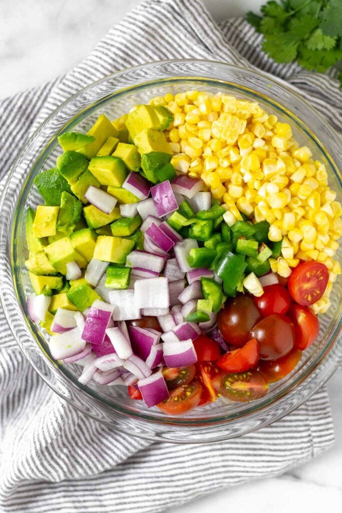 Large glass bowl of corn kernels, diced tomatoes, diced red onion, diced avocado, and diced jalapeños. The bowl is sitting on a towel and next to it is a bunch of cilantro.