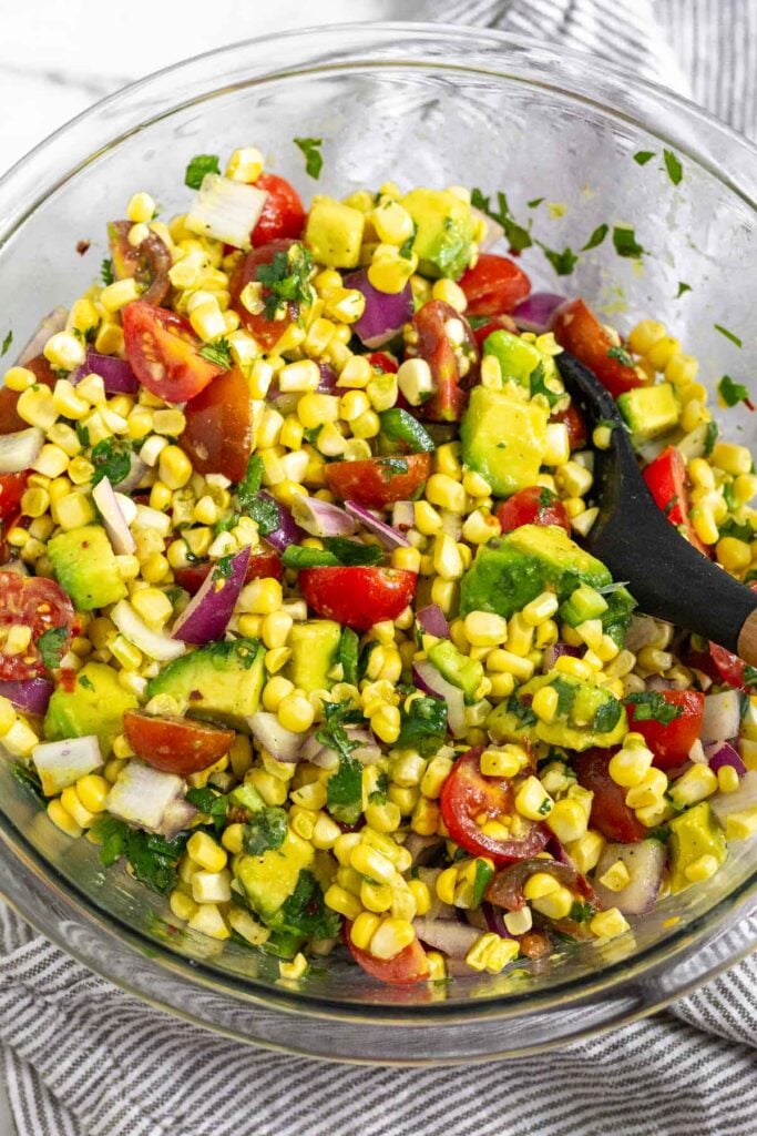 Mexican corn salad with tomato, avocado, red onion, and cilantro in a large glass bowl with a spoon in it.