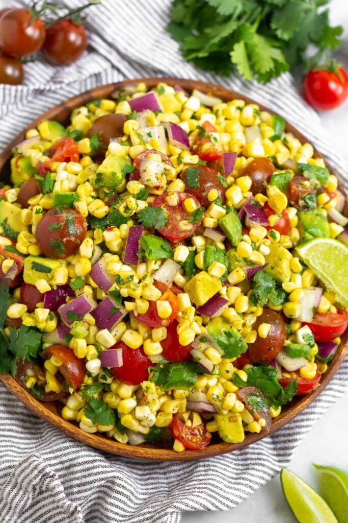 Tomato corn salad with avocado and cilantro in a large bowl sitting on a striped towel. Around it is some lime wedges, a bunch of cilantro, and some cherry tomatoes.