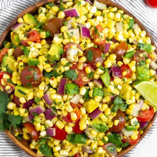 Summer corn salad in a large bowl with fresh corn kernels, diced tomato, diced avocado, diced red onion, and cilantro. The bowl is sitting on a striped towel and around it is some lime wedges, a bunch of cilantro, and some cherry tomatoes.