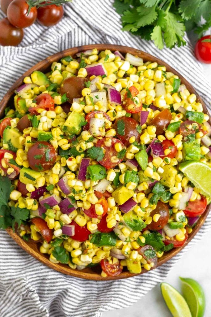 Summer corn salad in a large bowl with fresh corn kernels, diced tomato, diced avocado, diced red onion, and cilantro. The bowl is sitting on a striped towel and around it is some lime wedges, a bunch of cilantro, and some cherry tomatoes.