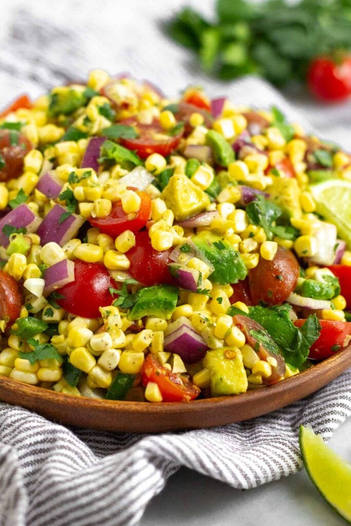 Corn avocado tomato salad in a large bowl garnished with avocado. The bowl is on a striped towel with a lime wedge in front of it and some cherry tomatoes and cilantro behind it.