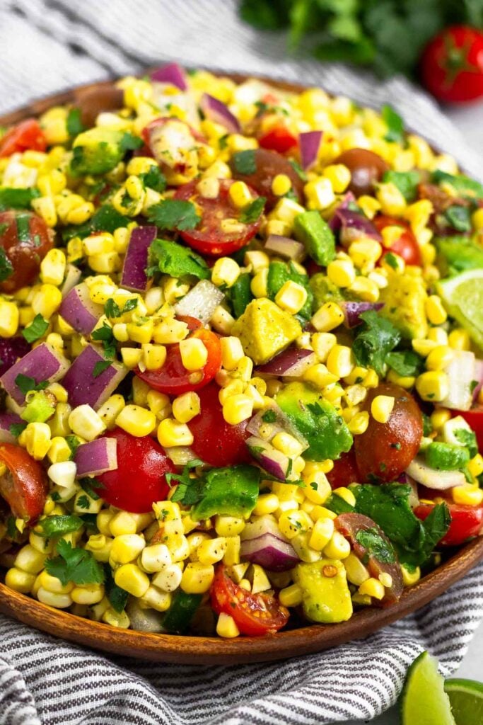 Large plate of corn salad with tomatoes, avocado, red onion, and cilantro. The plate is sitting on a striped towel with some cherry tomatoes, cilantro, and lime wedges.