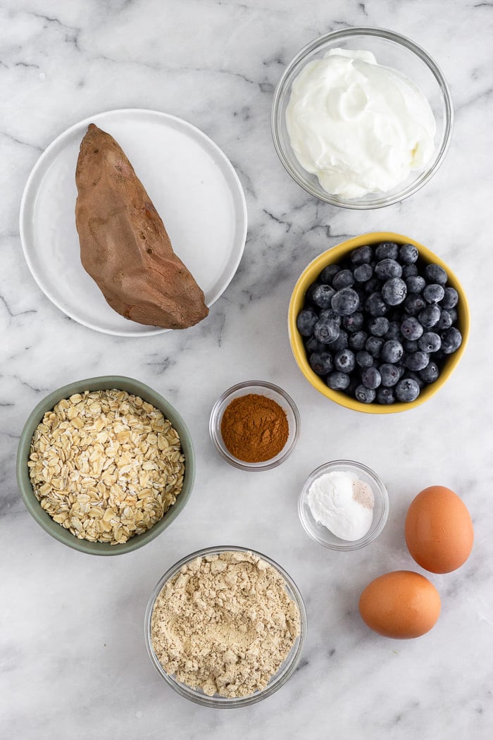 White marble counter top with a cooked sweet potato, greek yogurt, blueberries, cinnamon, eggs, oats, protein powder, and baking powder and soda.