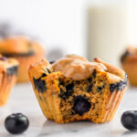 Healthy sweet potato blueberry protein muffin with a bit out of it and topped with peanut butter. There are some blueberries around it and more muffins and a jar of milk behind it.