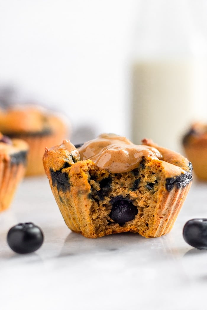 Healthy sweet potato blueberry protein muffin with a bit out of it and topped with peanut butter. There are some blueberries around it and more muffins and a jar of milk behind it.