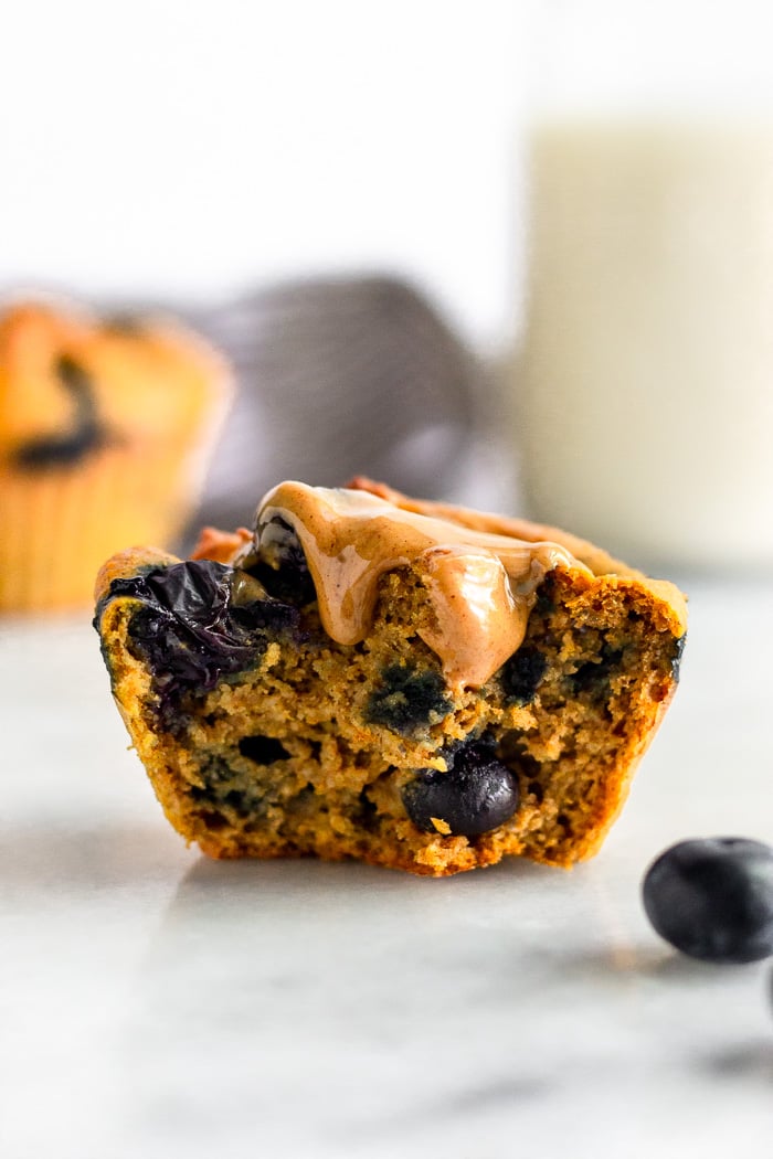 Half a sweet potato blueberry protein muffin standing up topped with peanut butter. Another muffin is behind it with a jar of milk.
