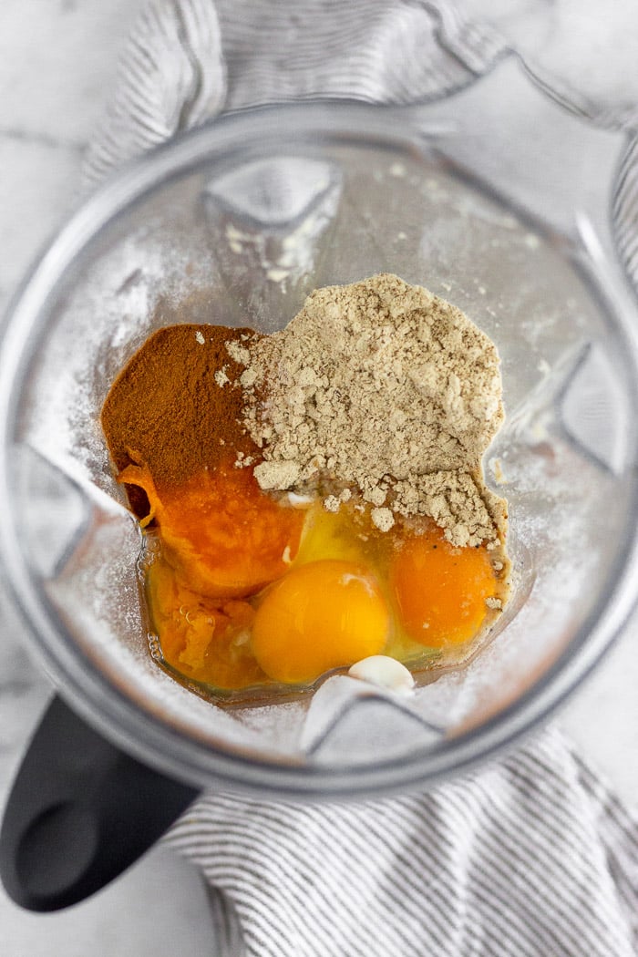 Blender filled with oat flours, sweet potato, cinnamon, protein powder, and eggs not yet blended.