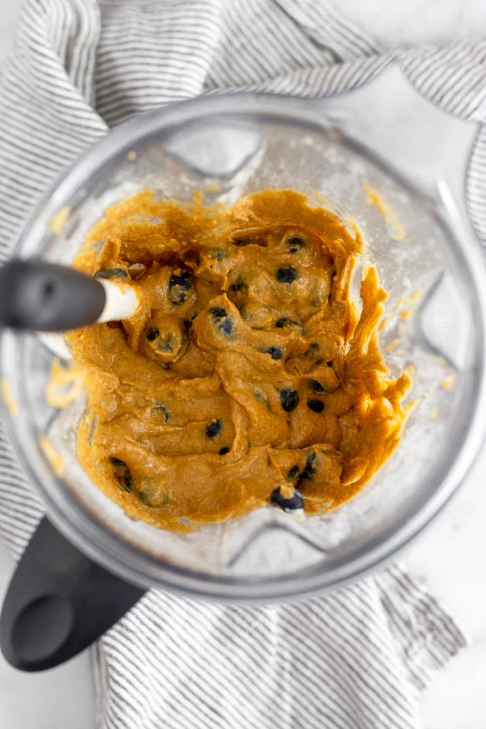 Blender filled with the mixture for sweet potato protein muffins with blueberries stirred in and a spatula sticking out.