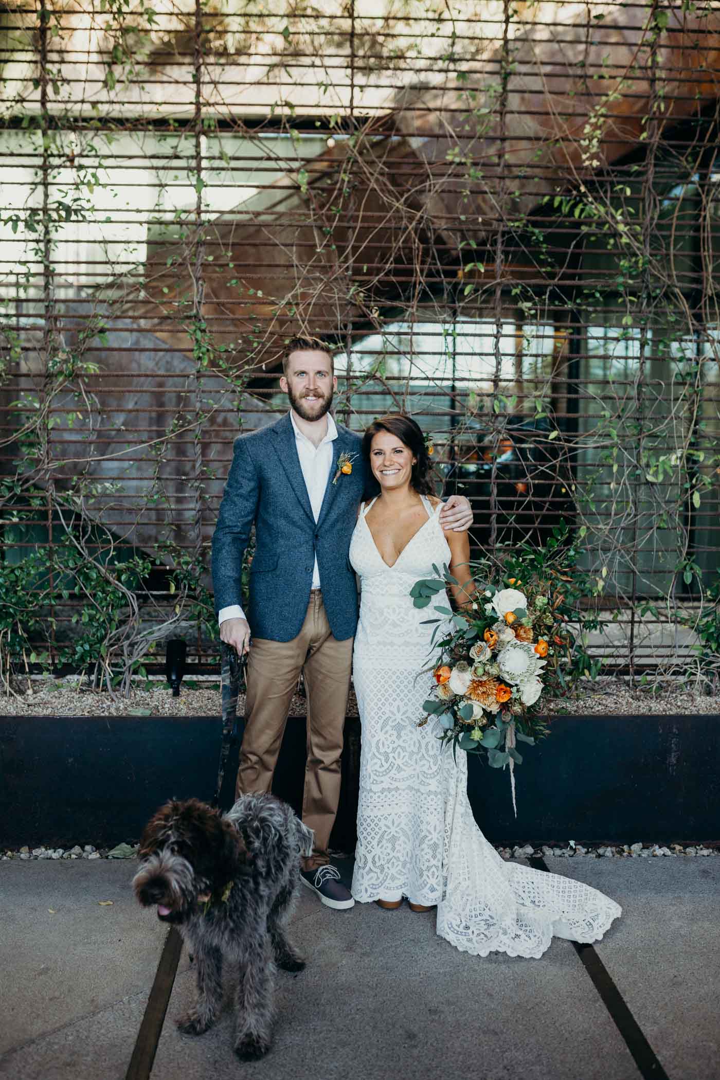 Bride and groom and their dog taking a picture in front of some greenery before they get married. Bride is wearing boho dress and the groom is wearing khakis and a jacket.