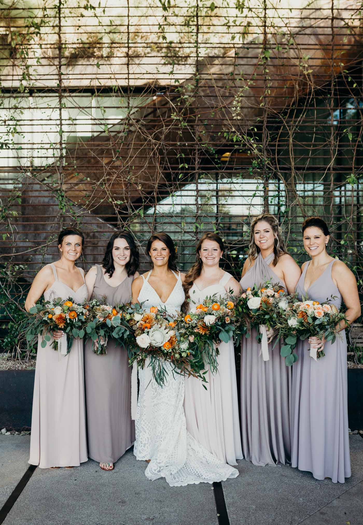 The bride and bridesmaids. The bride has a lace boho dress and the bridesmaids are all wearing different shares of tan. They are all holding flowers with greenery in it.