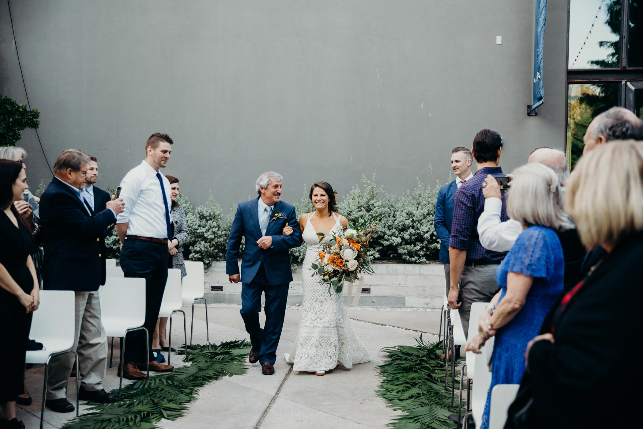Dad and his daughter walking down the aisle in her wedding. She is wearing a boho dress and holding flowers and palm leaves line the aisle.