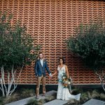 Bride and groom holding hands in front of a patterned wall with trees around them. The bride is wearing a boho dress and the groom is wearing khakis, a white shirt, and a blue jacket.