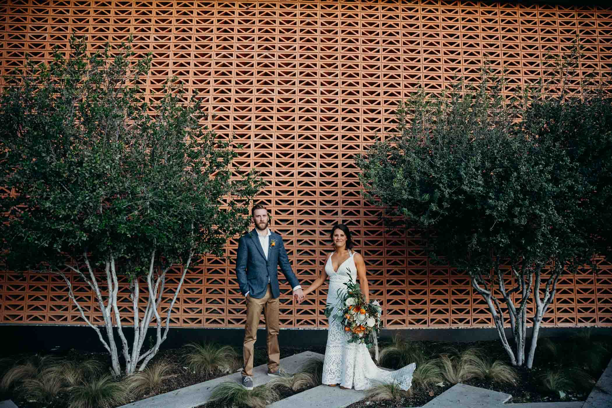 Bride and groom holding hands in front of a patterned wall with trees around them. The bride is wearing a boho dress and the groom is wearing khakis, a white shirt, and a blue jacket.