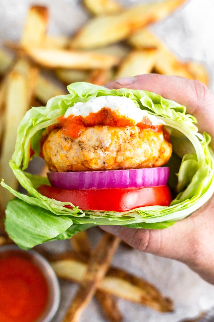 Person holding a buffalo chicken burger topped with buffalo sauce and blue cheese and wrapped in lettuce. Behind it is a pile of fries.