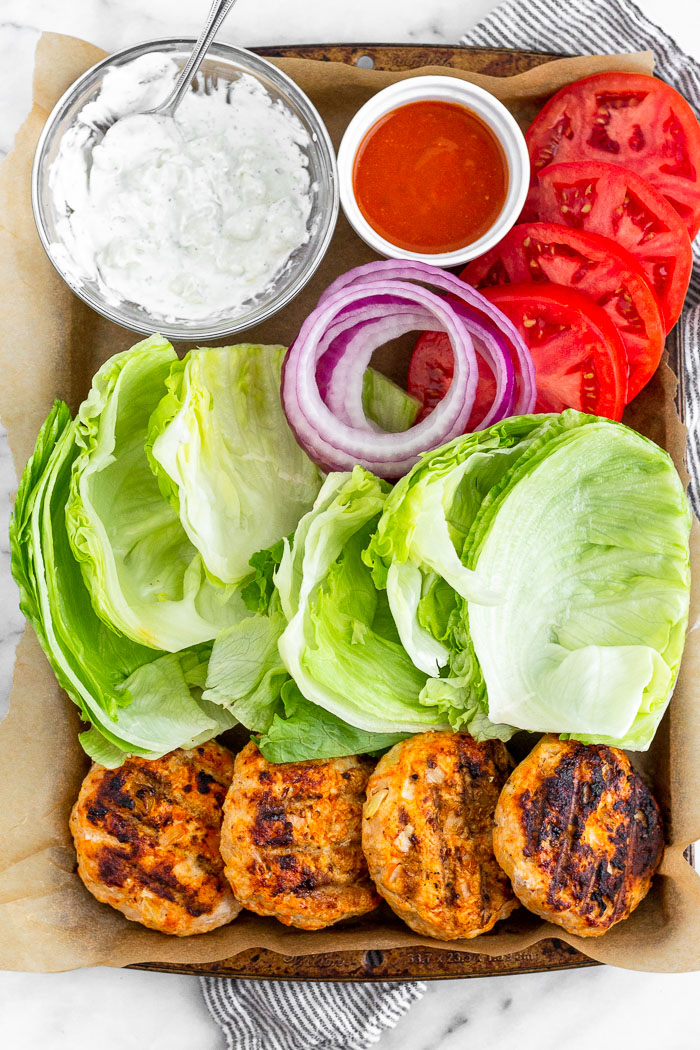 Platter of buffalo chicken burgers, lettuce leaves, sliced tomatoes and red onion, small bowl of buffalo sauce, and a bowl of blue cheese dressing.