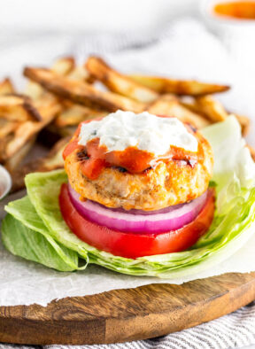 Buffalo chicken burger in a lettuce wrap topped with buffalo sauce and blue cheese dressing. Behind it is a pile of French fries.