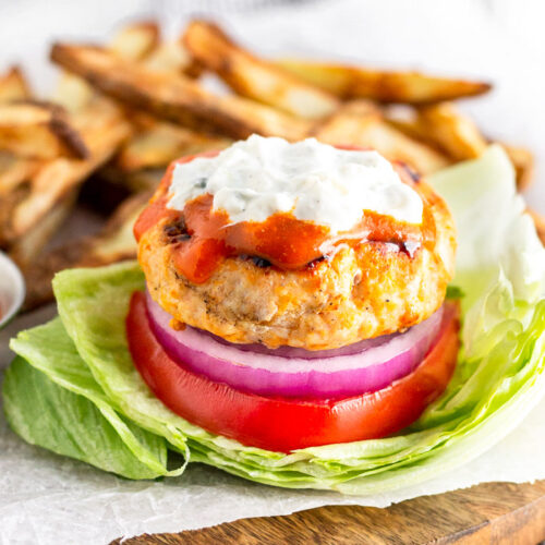 Buffalo chicken burger in a lettuce wrap topped with buffalo sauce and blue cheese dressing. Behind it is a pile of French fries.