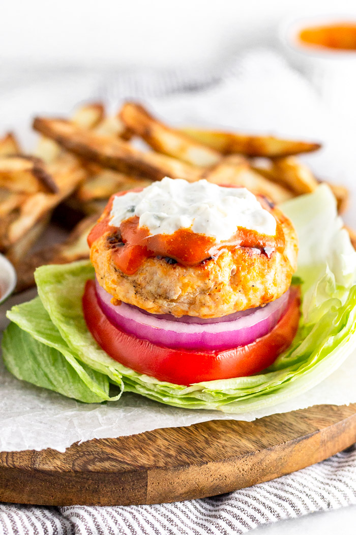 Buffalo chicken burger in a lettuce wrap topped with buffalo sauce and blue cheese dressing. Behind it is a pile of French fries.
