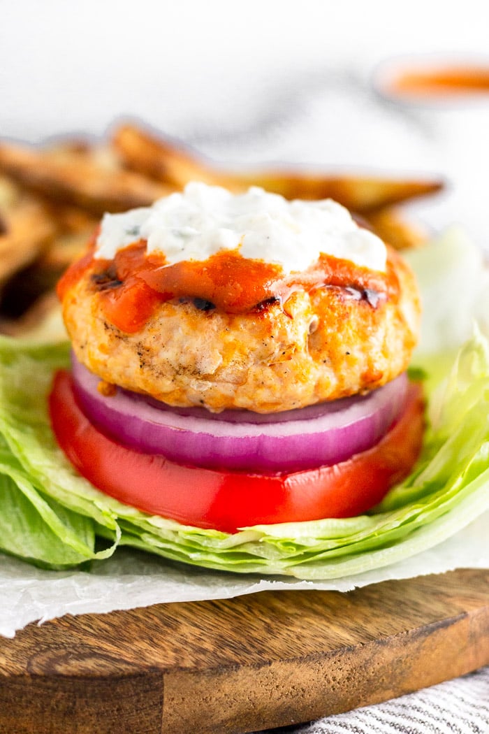 Grilled buffalo chicken burger topped with buffalo sauce and blue cheese dressing sitting on a lettuce bun with sliced tomato and onion. It is sitting on a piece of paper on top of a cutting board.