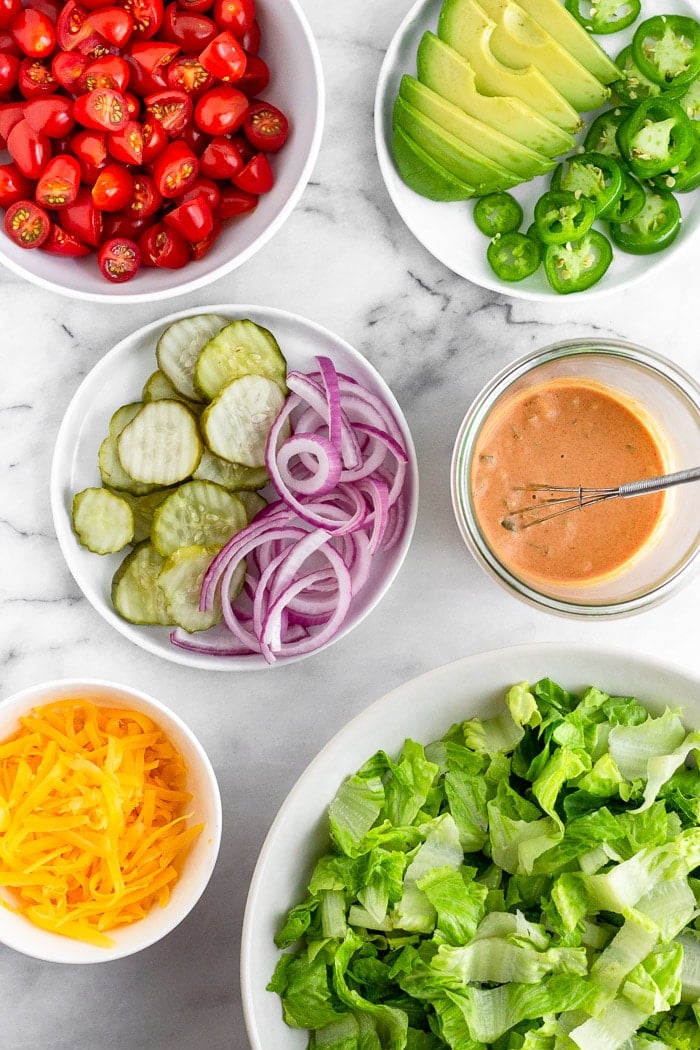 White counter filled with a bowl of lettuce, bowl of shredded cheese, plate of pickles and onions, jar of special sauce, plate of jalapeños and avocado, and a bowl of chopped tomatoes.
