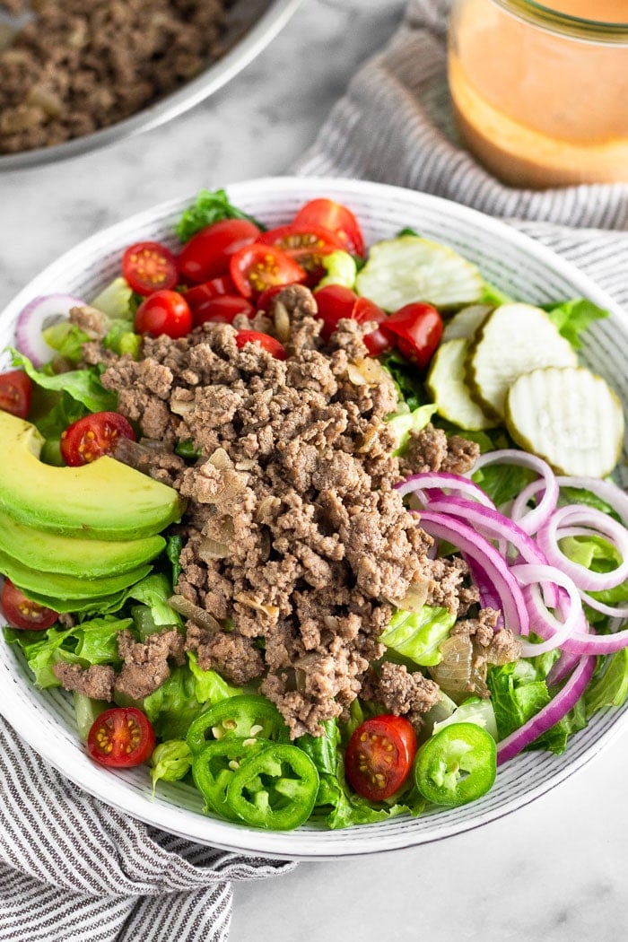 Paleo burger bowl with lettuce, tomatoes, pickles, onions, jalapeños, avocado, and ground beef. Behind it is a pan of ground beef and a jar of special sauce.