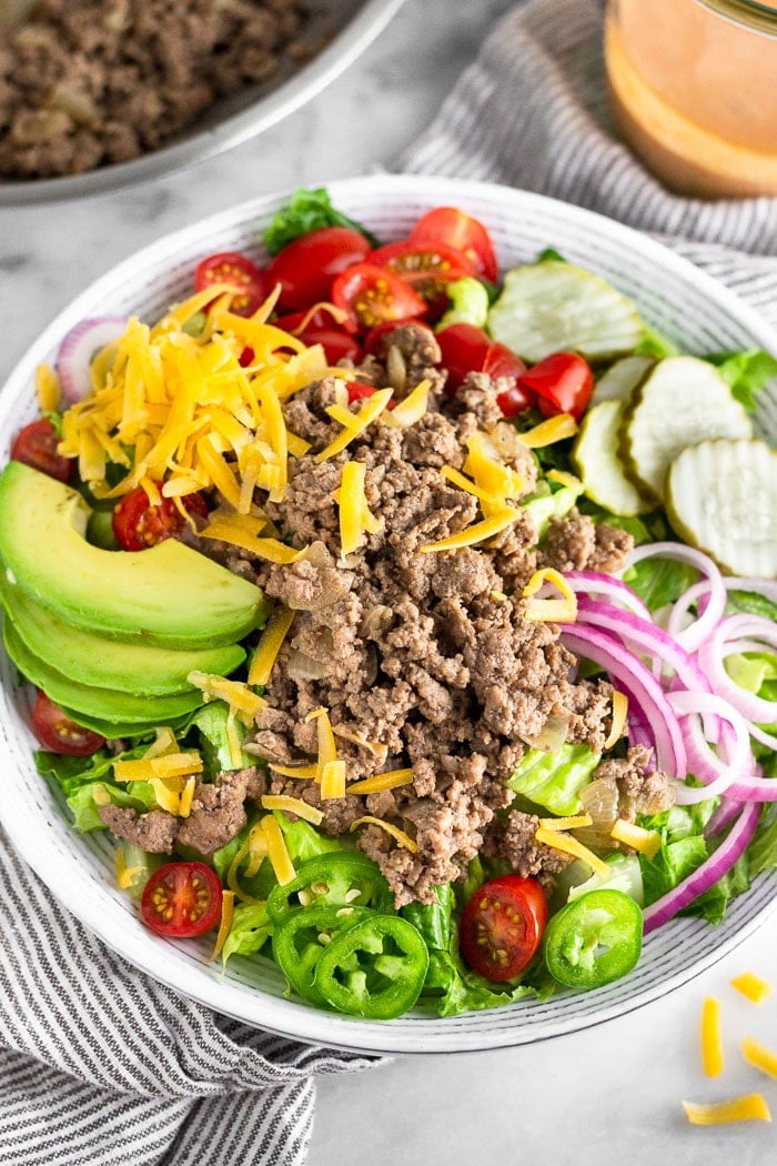 Cheeseburger salad with lettuce, tomatoes, pickles, onions, jalapeños, avocado, and ground beef. Behind it is a pan of ground beef and a jar of special sauce.