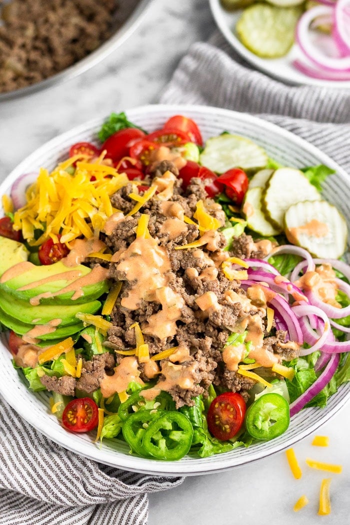 Burger salad with lettuce, tomato, onion. pickles, avocado, shredded cheese, jalapeños, and special sauce dressing. Behind it is a pan of ground beef and a plate of more toppings.